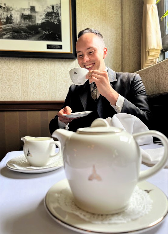 A 20-35 year old man in a dark suit is smiling and sipping tea in the Legislative Assembly dining room.