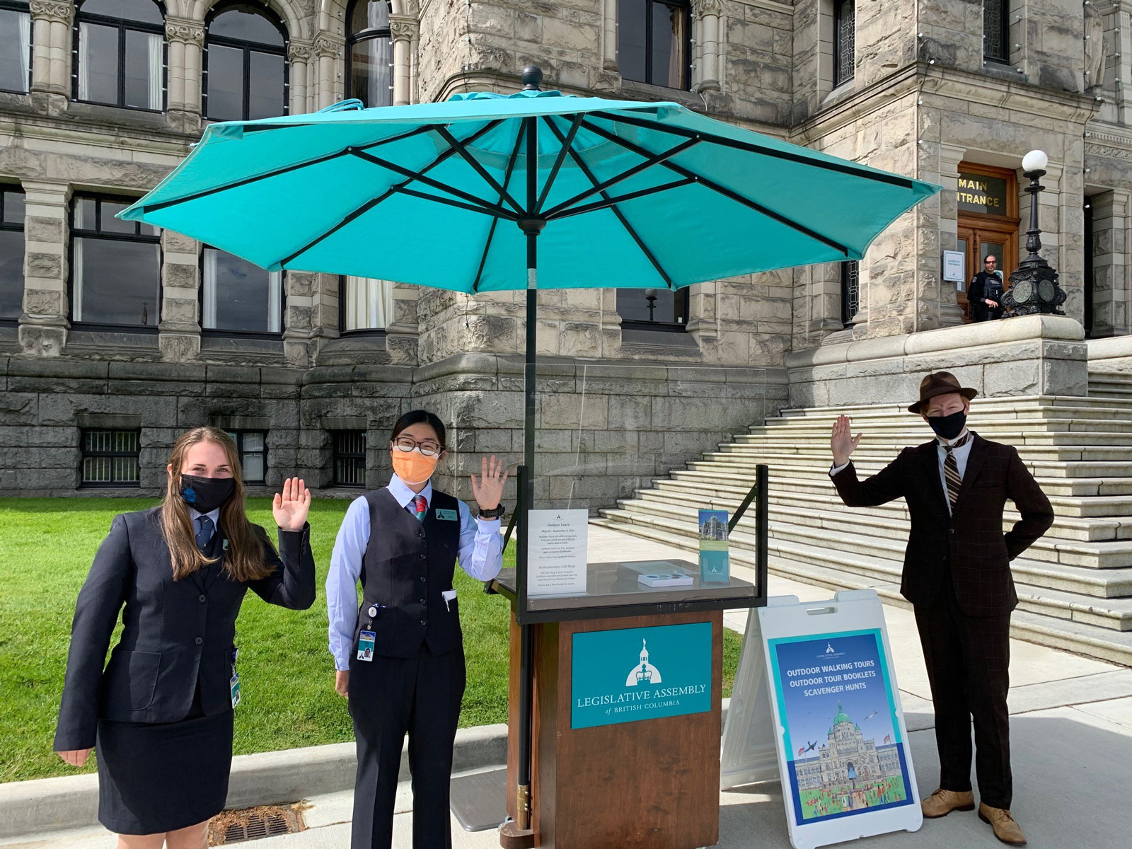 3 Legislative Assembly summer tour guides waving standing beside the tour guide kiosk beside the Legislative Assembly steps to the front door, with a turquoise umbrella overhead.